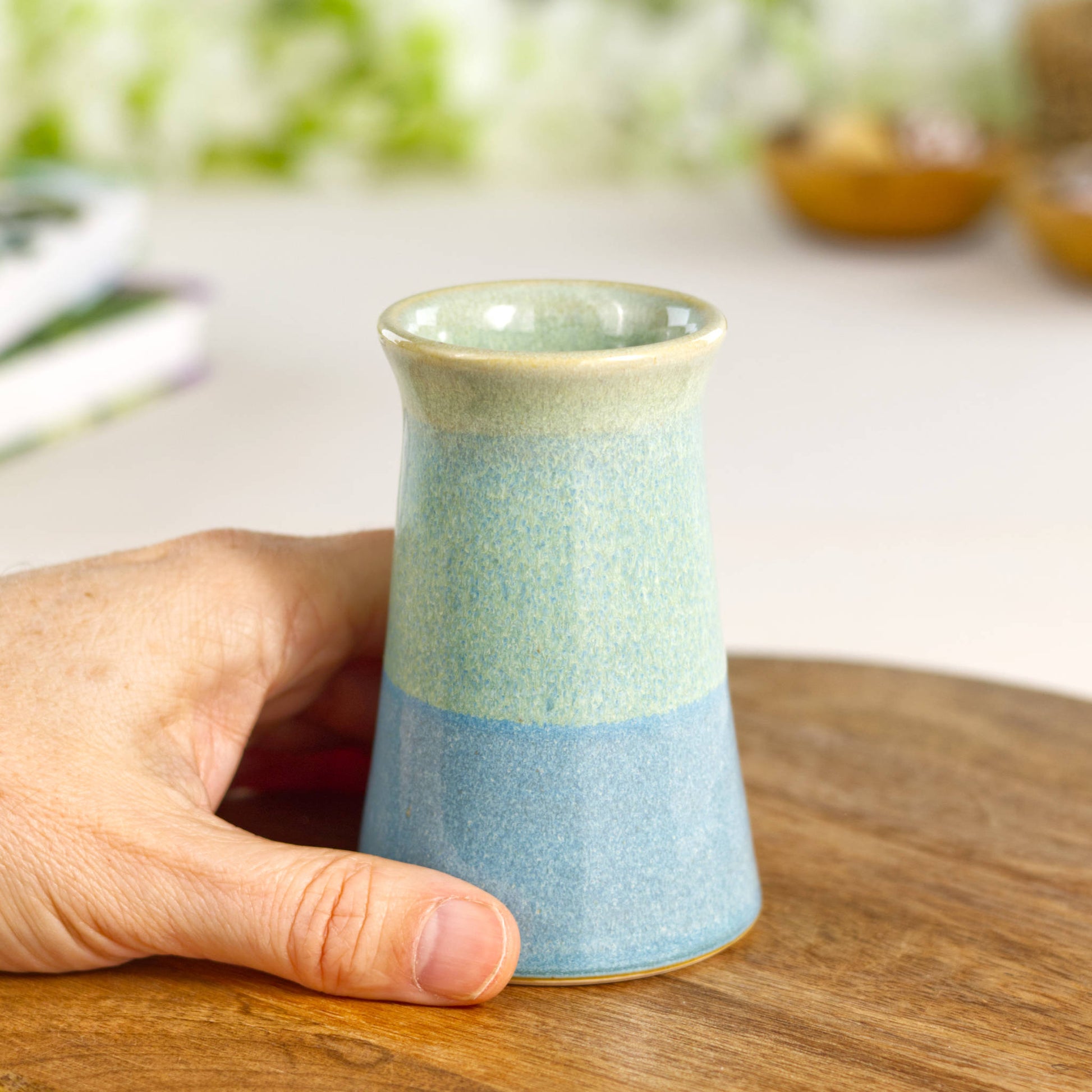Small handcrafted bud vase in coastal blue and pale green tones, a charming two-tone tapered design shown in hand to highlight its petite proportions and homely appeal.