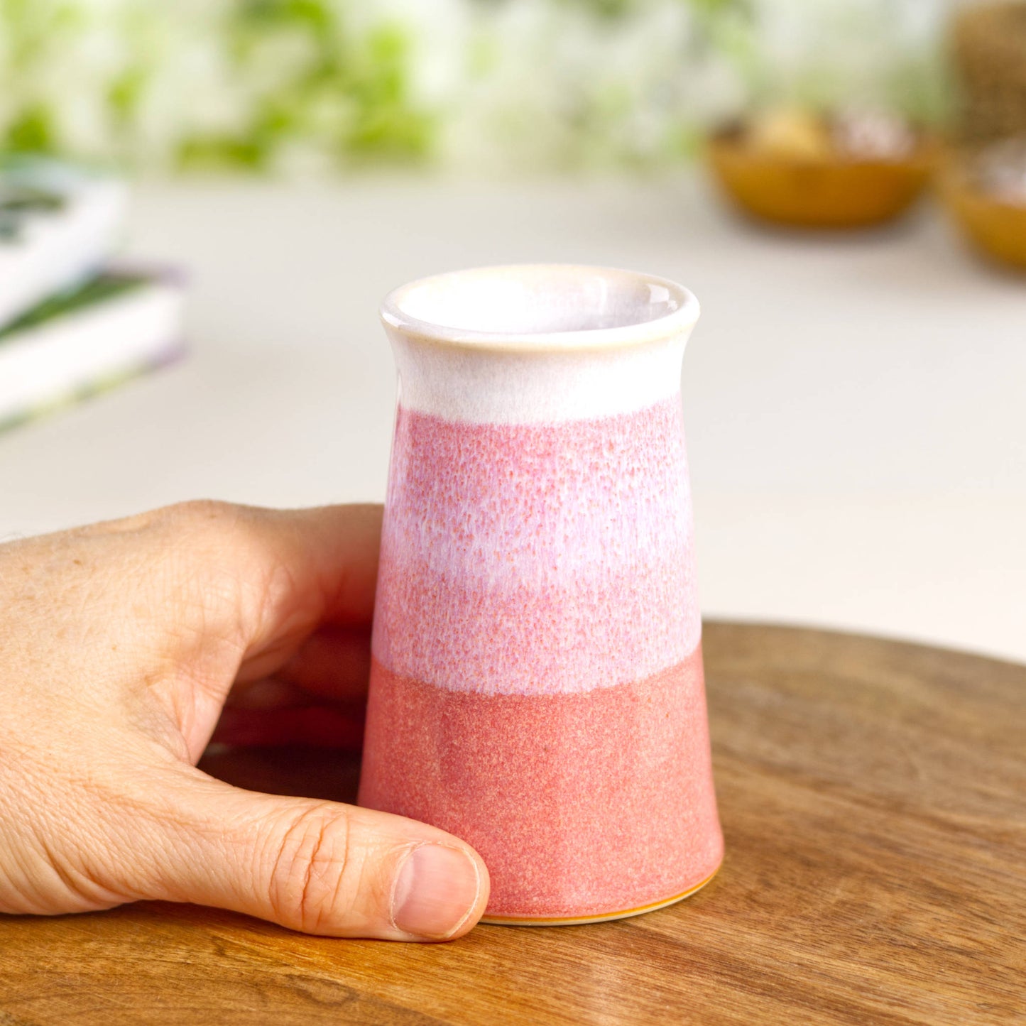 Hand holding a small coral pink and white conical bud vase, handmade wheel-thrown unique British pottery shown for scale.