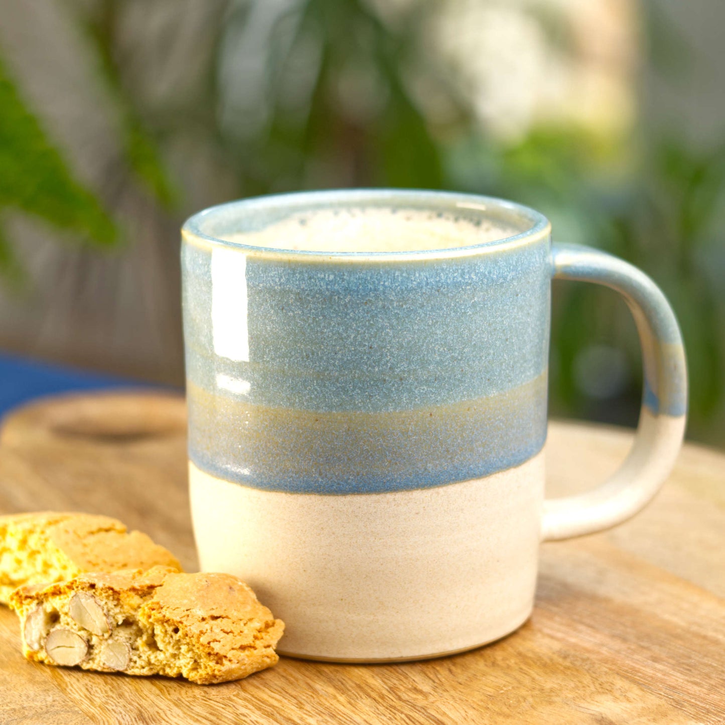 Coastal Blue and Sand handmade ceramic mug filled with frothy coffee on a wooden board with biscotti, wheel-thrown stoneware from my Sussex studio