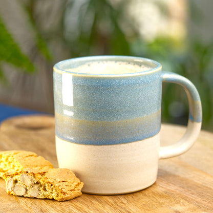 Coastal Blue and Sand handmade ceramic mug filled with frothy coffee on a wooden board with biscotti, wheel-thrown stoneware from my Sussex studio
